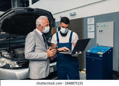 Car Mechanic Talking With Senior Business Man Costumer During Periodic Car Condition Check. They Are Wearing Protective Face Mask Against Virus Pandemic.