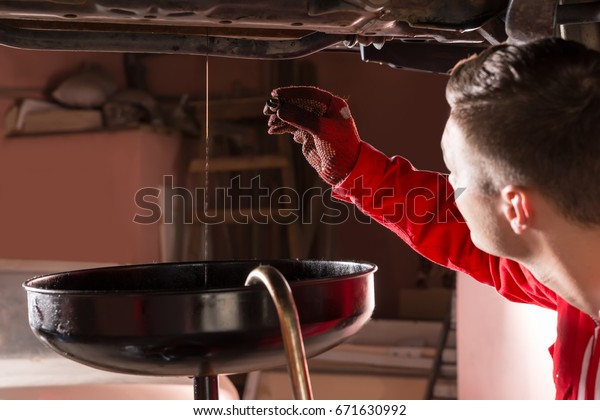 Car mechanic in uniform watching how oil\
flows out and changing motor oil in automobile engine at\
maintenance repair service station in a car\
workshop