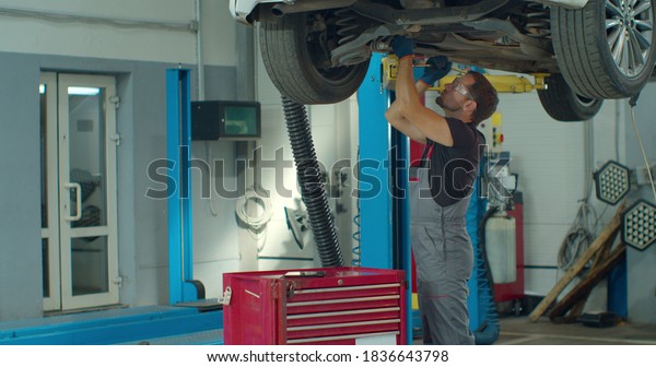 Car mechanic is working under a vehicle on a\
lift in service. Repairman is using ratchet. Specialist is wearing\
safety glasses.