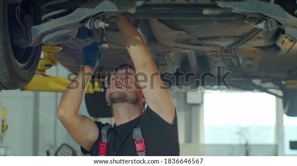 Car mechanic is working under a vehicle on a\
lift in service. Repairman is using ratchet. Specialist is wearing\
safety glasses.