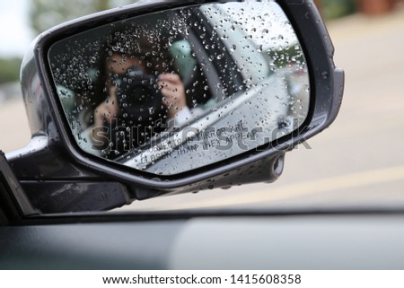 Eine Frau in einer mit großem Ausschnitt sitzt in ihrem Auto auf der rechten Seite vorne und schminkt sich, legt Schmuck an. Man kann sie durch die Reflexion in der Windschutzscheibe nicht erkennen