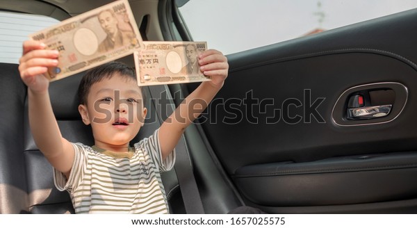 Car and money. Happy little boy sitting in car and\
holding Japanese money \