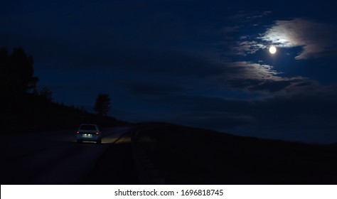 Car Moves At Night On A Road Under A Cloudy Sky Lit By The Moon