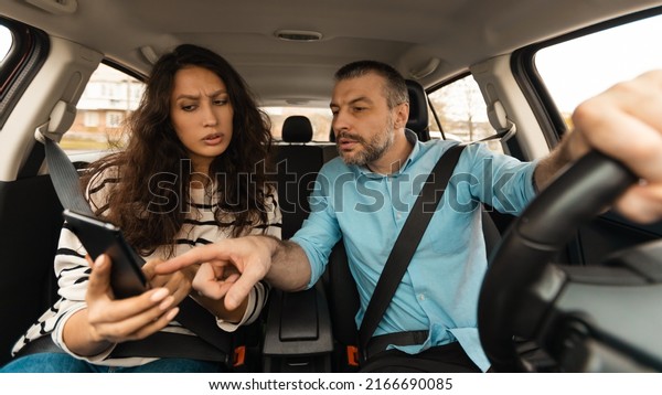 Car\
Navigation Problem. Confused Couple Riding Auto, Man Driving And\
Pointing Finger At Smartphone Using Mobile Application Sitting In\
Driver\'s Seat Indoor. Traffic App Concept.\
Panorama