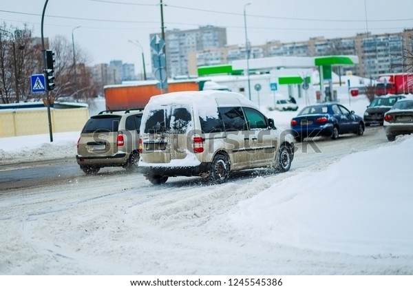 car on cleared road\
after heavy snowfall and snowstorms in city. Traffic lights hang on\
wire above road. included headlights at car in winter. Car with\
snow on roof.