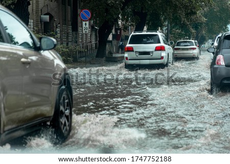 Вriving car on flooded road during flood caused by torrential rains. Cars float on water, flooding streets. Splash on car. Flooded city road with large puddle.  Flooding after heavy rains at city