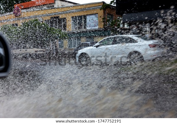 Вriving car on flooded road during flood caused by\
torrential rains. Cars float on water, flooding streets. Splash on\
car. Flooded city road with large puddle.  Flooding after heavy\
rains at city