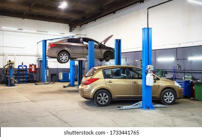 Car On Lift In Mechanic Shop Or Garage, Interior Of Auto Repair Workshop, Vehicles Inside Maintenance. Auto Hoist In Small Service Station. Concept Of Workplace And Vehicle Business.