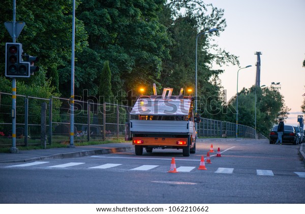 Car on side of road with warning lights. Road\
construction works. Traffic line painting. Painting white street\
lines on pedestrian crossing. Road cones with orange, white stripes\
standing on asphalt