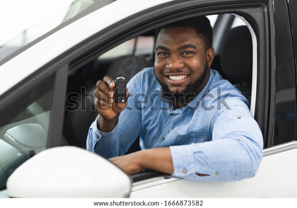 Car Owner. Joyful\
Afro Man Showing Automobile Key Sitting In New Auto In Dealership\
Showroom. Selective Focus