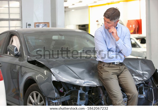 Car owner sitting on the bonnet of a damaged car and\
looking at it.
