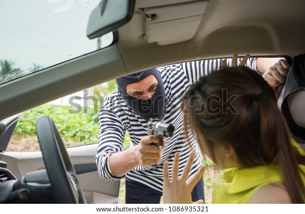 Car owner threatened by masked gun robber during\
day time, on raining day. Gangster criminal cover face with black\
mask pointing gun at young girl. Property & life Insurance and\
Safety concept.