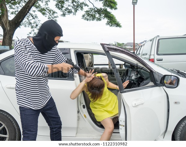 Car owner threatened by masked gun robber during\
day time, on raining day. Gangster criminal cover face with black\
mask pointing gun at young girl. Property & life Insurance and\
Safety concept.