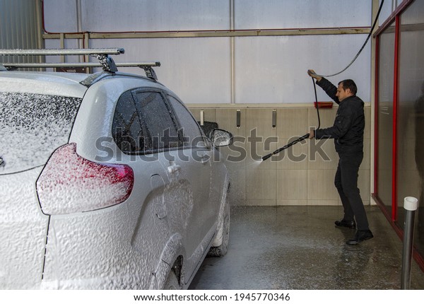 Car owner\
washes the car under high pressure with foam and water in a car\
wash with a serious face. Car care\
concept.