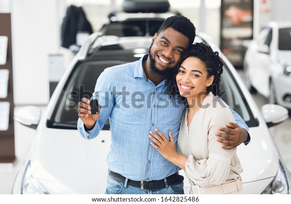 Car Owners.
Happy African American Couple Showing Automobile Key Embracing
Standing In Dealership
Showroom.