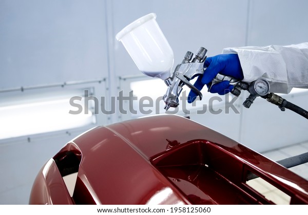 Car\
painter in protective clothes and mask painting automobile bumper\
with metallic paint and varnish in chamber\
workshop.