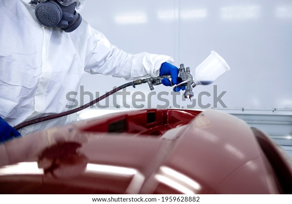 Car\
painter in protective clothes and mask painting automobile bumper\
with metallic paint and varnish in chamber\
workshop.