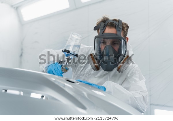 Car painter in protective\
clothes and mask painting and varnish automobile bumper in chamber\
