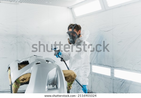 Car painter\
in a protective suit and mask varnishes a painted bumper of a\
vehicle while working in a painting\
booth