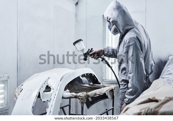 Car painting and automobile repair service.\
Auto mechanic in white overalls paints car with airbrush pulverizer\
in paint chamber\
