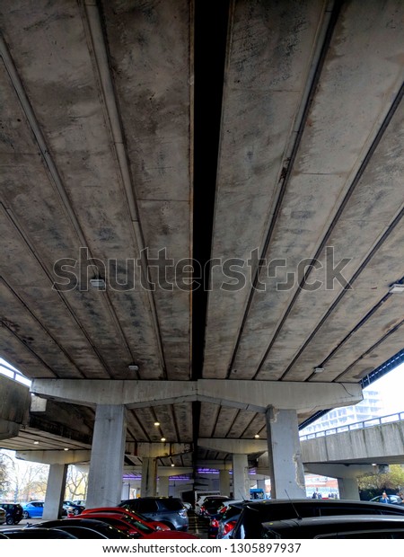 A car park\
underneath a modern, concrete, Brutalist, road bridge (flyover),\
with the tops of cars showing
