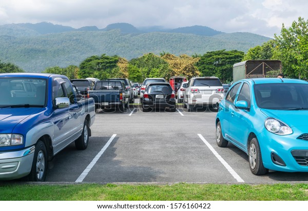 Car parked in asphalt parking lot. Trees,\
mountain, blue sky background, empty space parking. Outdoor parking\
lot with green environment. Travel transportation technology with\
nature concept\
