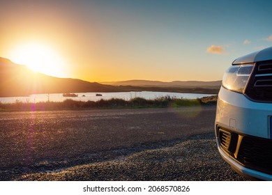 Car Parked By A Small Lake With Blue Water And Wild Fields. Beautiful Sun Rise Scene In Connemara, County Galway, Ireland. Warm And Cool Tones. Haze Over Mountains In The Background.