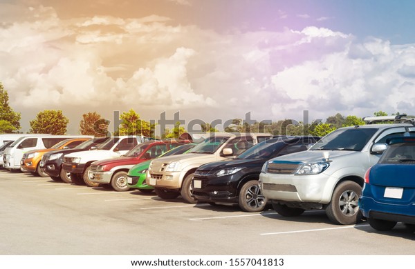 Car parked in large asphalt parking lot in a\
row with white cloud and blue sky background. Outdoor parking lot\
with fresh ozone and green environment of travel transportation\
technology concept\
