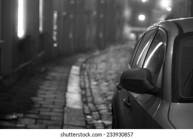 Car Parked In Narrow Old Street With Cobblestones At Night. Black And White.