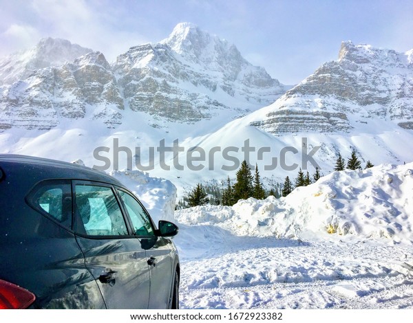 A car parked overlooking incredible view of a dark\
green forest in the foreground and snow covered mountains peaks in\
the background, along the icefield parkway in the Rocky Mountains\
Alberta, Canada