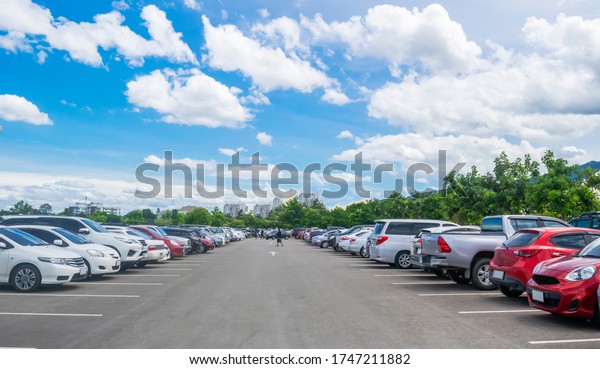 Car parking in large asphalt parking lot with\
trees, white cloud and blue sky background. Outdoor parking lot\
with fresh ozone and green environment of transportation and modern\
technology concept\
