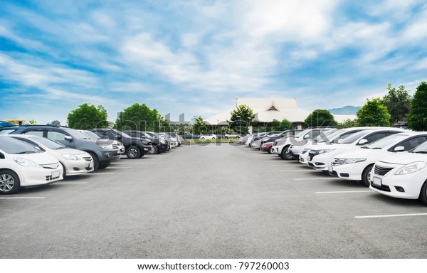 Car\
parking in large asphalt parking lot with trees, white cloud and\
blue sky background in front of hall building. Outdoor parking lot\
with fresh ozone and green environment\
concept