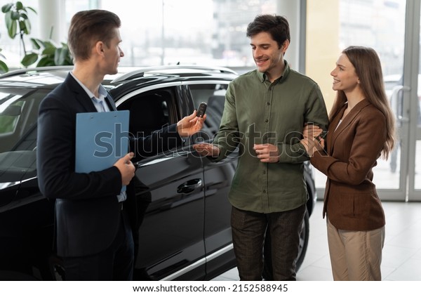 Car purchase or lease\
concept. Cheerful young couple taking car key from auto salesman at\
modern dealership. Millennial spouses buying new automobile at\
showroom store