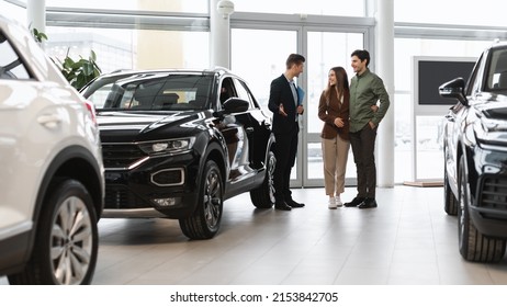 Car purchase or rental. Positive young spouses speaking to salesman about buying new auto at dealership, panorama. Successful millennial manager showing clients choice of vehicles at salon - Powered by Shutterstock