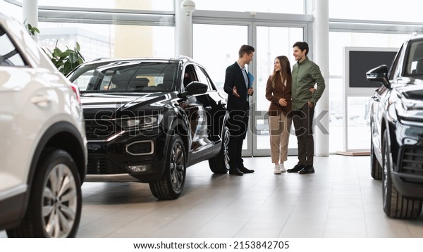 Car purchase or rental. Positive young spouses\
speaking to salesman about buying new auto at dealership, panorama.\
Successful millennial manager showing clients choice of vehicles at\
salon