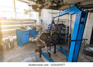 A Car Raised On A Rotary Lift For Repair And Under It A Detached Engine Suspended On A Blue Crane Near Workbench In Auto Service.