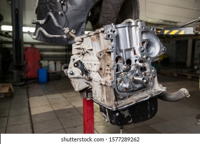 A Car Raised On A Rotary Lift For Overhaul Repair And Under It A Detached Bottom Engine Part Suspended On A Red Crane Near Workbench In Vehicle Workshop. Auto Service Industry.