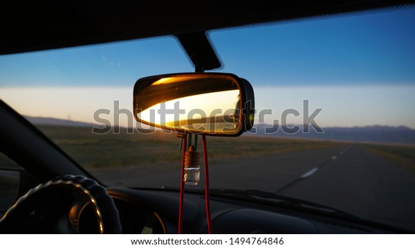 Car rear view mirror. Displays orange dawn.\
Visible road, hills and steppe. Hang key chains. The car goes fast.\
Kazakhstan road, near\
Almaty.