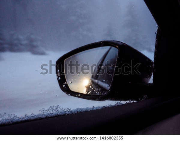 Car rear view mirror\
of driver reflection with snowy road and car following on the\
forest winter road