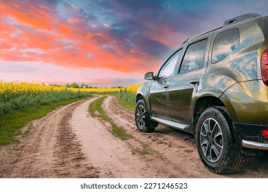 Car Renault Duster Or Dacia Duster Suv In Summer Rapeseed Field Countryside Landscape On Background Bright Orange Dramatic Sky. Summertime Flowering Canola, Rapeseed, Oilseed Field Meadow Grass.