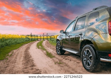 Car Renault Duster Or Dacia Duster Suv In Summer Rapeseed Field Countryside Landscape On Background Bright Orange Dramatic Sky. Summertime Flowering Canola, Rapeseed, Oilseed Field Meadow Grass.
