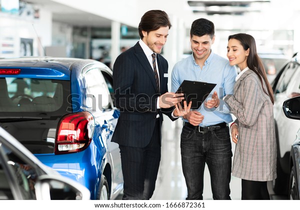 Car Rent.\
Handsome Salesman Offering Couple Automobile, Showing Clipboard,\
Standing In Auto Rental\
Office
