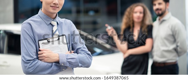 Car rental banner size concept. Asian officer\
male staff stands and holding rental agreement document in front.\
Couple Caucasian man and woman holding remote car key near vehicle\
behind in background.