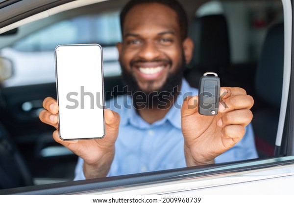 Car Renting App. Happy Black Man Showing Key And\
Smartphone With Blank White Screen Through Window While Sitting In\
His Automobile In Dealership Center Or Rental Office, Closeup Shot,\
Mockup