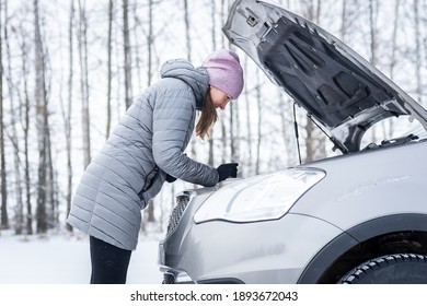 Car Repair On The Road In Winter. Young Girl Is Trying To Fix A Car Breakdown Under The Soot On The Road. Woodsroadside Assistance Car