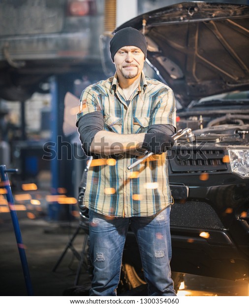 Car repair service. Brutal mechanic man\
stands next to a car and holding a\
wrench