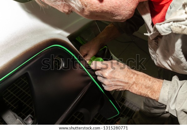 A car repair worker during work pastes with an\
adhesive tape a part of the body element of white color in order\
not to fill it with paint during painting in a workshop on repair\
of vehicles