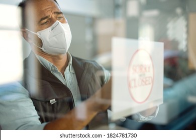 Car Repairman With Protective Face Mask Hanging Closed Sign While Going Out Of Business Due To COVID-19 Pandemic. The View Is Through The Glass. 