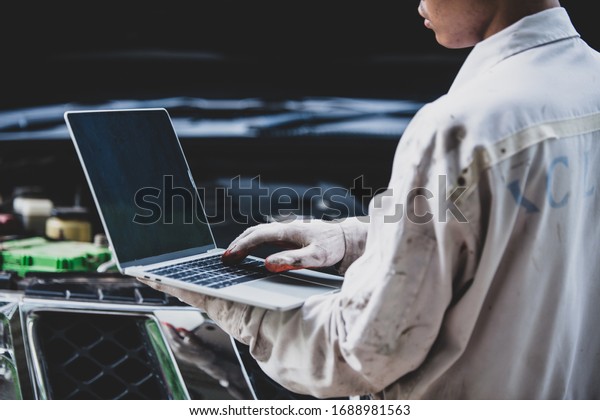 Car repairman wearing a white uniform standing and\
holding a wrench that is an essential tool for a mechanic with\
laptop checking engine