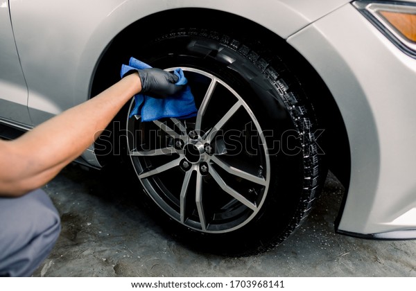 Car rims\
cleaning, car detailing wash concept. Cropped close up photo of\
male hand in black rubber glove with blue microfiber cloth washing\
car alloy wheel at car wash\
service